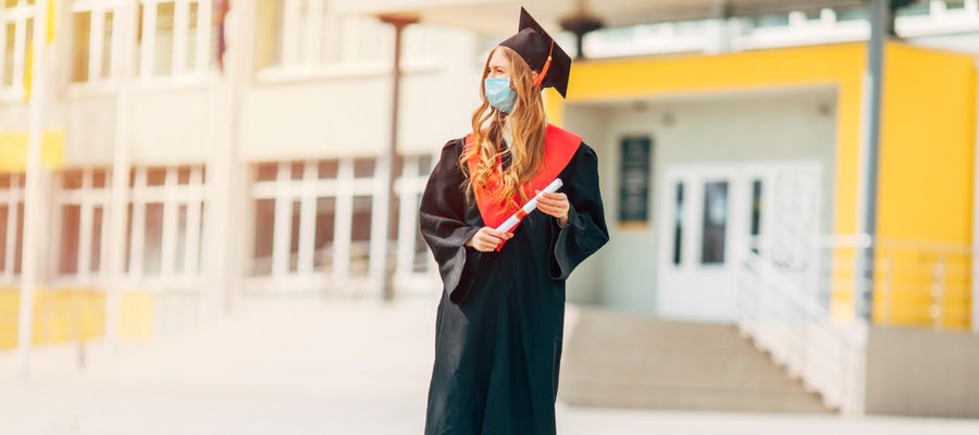 A lone woman wearing her graduation robe and cap and a facemask.